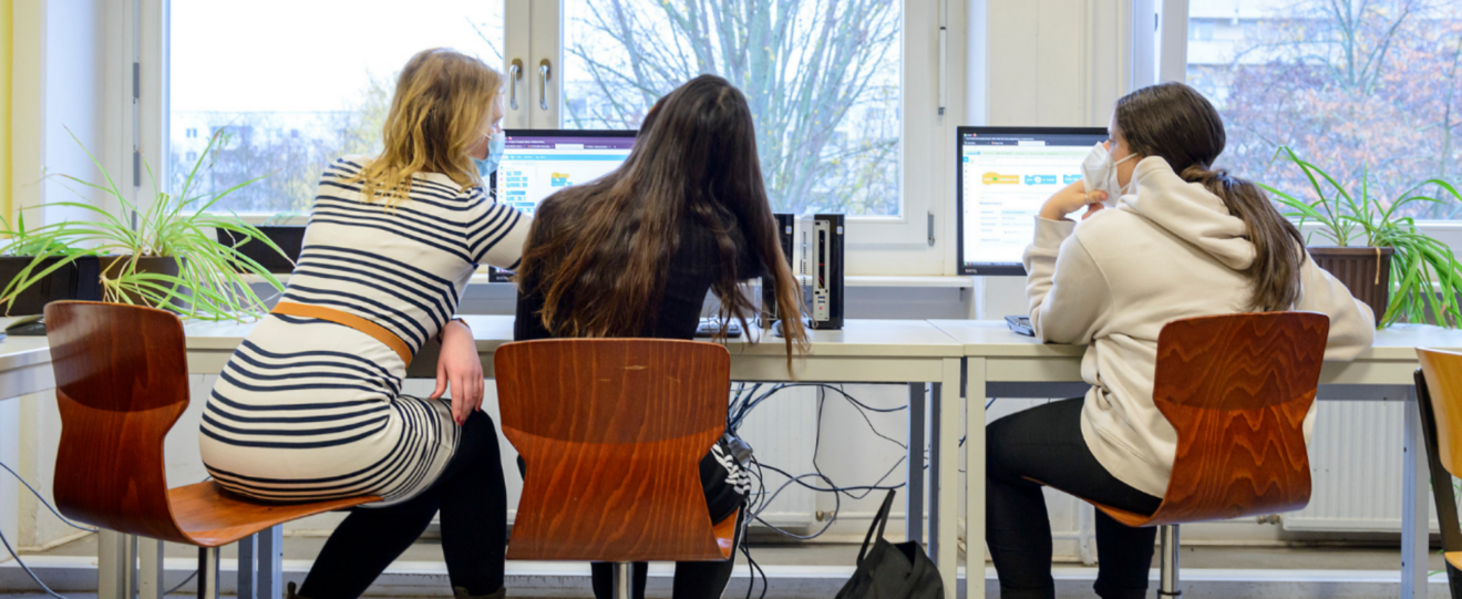 Schülerinnen sitzen an einem Schreibtisch, auf dem Tisch steht ein Computer, die Schülerinnen sitzen mit dem Rücken zur Kamera