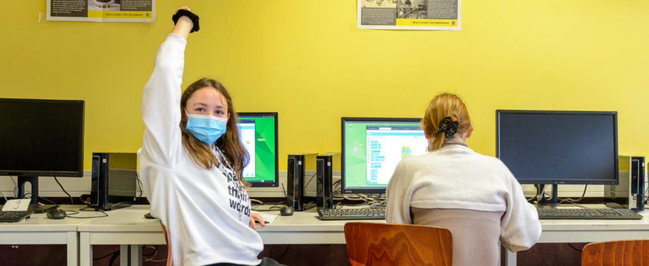 zwei Mädchen sitzen vor Computern, eines mit dem Rücken zur Kamera, das andere ist der Kamera zugewandt und meldet sich, sie trägt eine OP-Maske