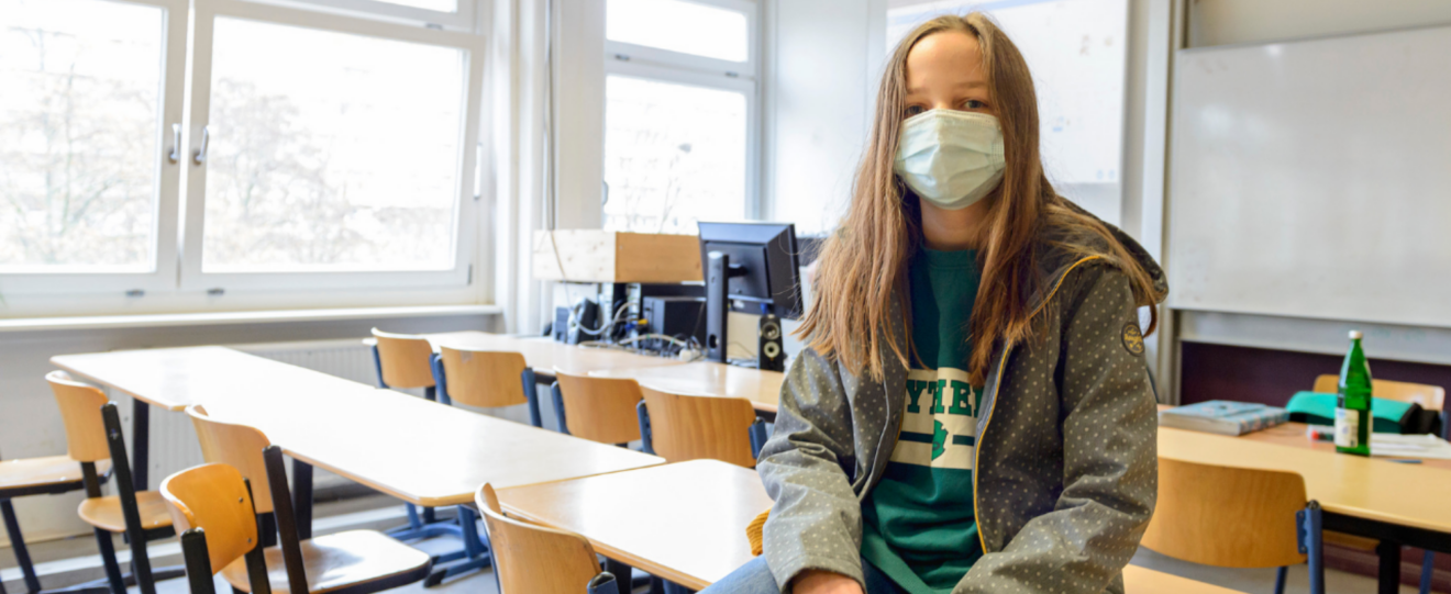 ein Mädchen sitzt in einem leeren Klassenzimmer auf dem Tisch, Porträt, sie trägt eine OP-Maske